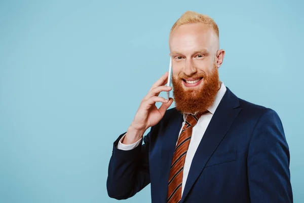 Sonriente hombre de negocios barbudo hablando en el teléfono inteligente, aislado en azul - foto de stock