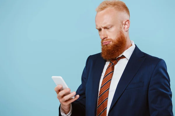 Hombre de negocios barbudo confundido hablando en el teléfono inteligente, aislado en azul - foto de stock