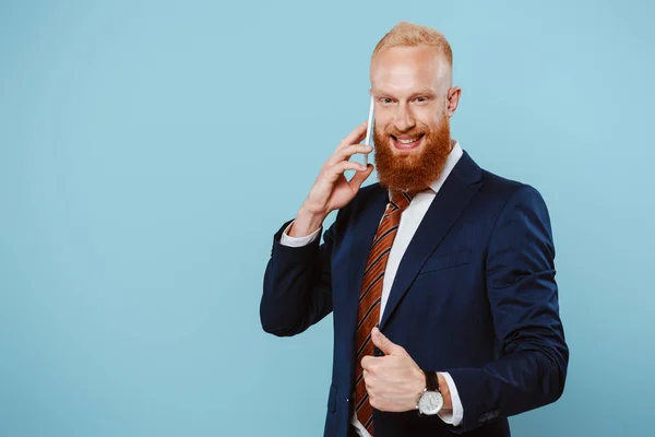 Sonriente hombre de negocios barbudo hablando en el teléfono inteligente y mostrando el pulgar hacia arriba, aislado en azul - foto de stock