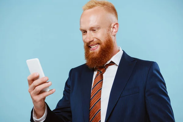Hombre de negocios barbudo feliz usando teléfono inteligente, aislado en azul - foto de stock