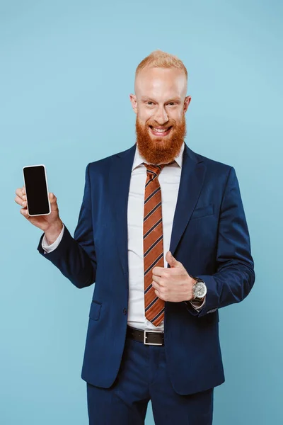 Sonriente hombre de negocios barbudo mostrando el pulgar hacia arriba y el teléfono inteligente con pantalla en blanco, aislado en azul - foto de stock