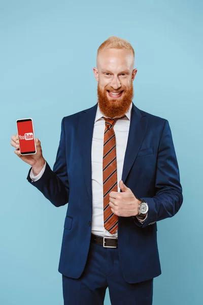 KYIV, UCRANIA - 27 de agosto de 2019: hombre de negocios barbudo sonriente mostrando el pulgar hacia arriba y el teléfono inteligente con la aplicación de youtube, aislado en azul - foto de stock