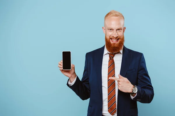 Uomo d'affari barbuto sorridente che punta a smartphone con schermo bianco, isolato su blu — Foto stock