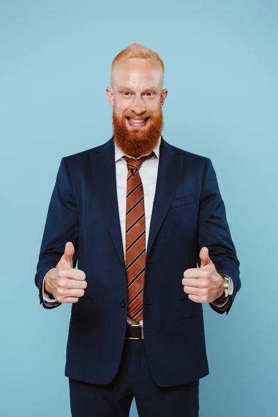 Sonriente hombre de negocios barbudo en traje mostrando los pulgares hacia arriba, aislado en azul - foto de stock