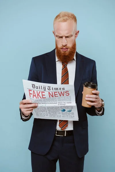Hombre de negocios barbudo serio leyendo el periódico con noticias falsas y sosteniendo café para llevar, aislado en azul - foto de stock