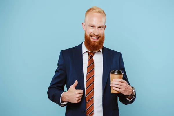 Allegro uomo d'affari barbuto in giacca e cravatta tenendo il caffè per andare e mostrando pollice in alto, isolato su blu — Foto stock