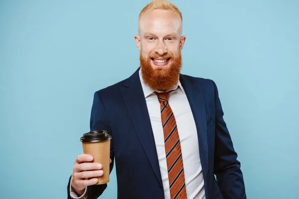 Sonriente hombre de negocios barbudo en traje sosteniendo café para llevar, aislado en azul - foto de stock