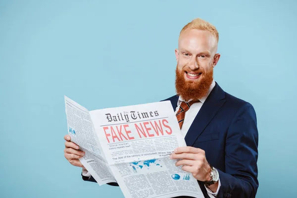 Sonriente hombre de negocios barbudo leyendo el periódico con noticias falsas, aislado en azul - foto de stock