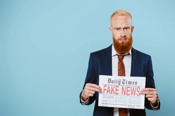 Preocupado hombre de negocios barbudo sosteniendo periódico con noticias falsas, aislado en azul - foto de stock