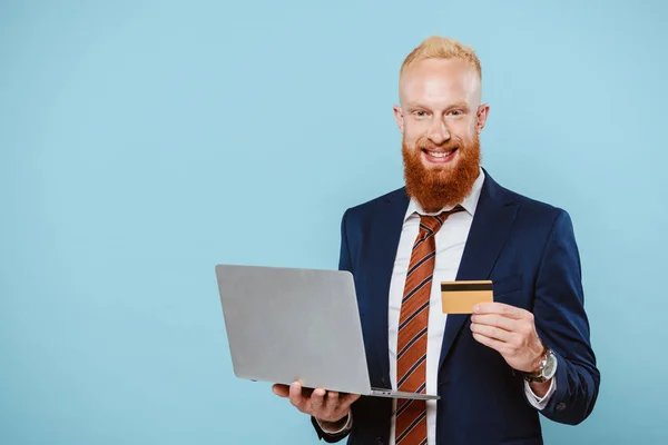 Happy bearded businessman in suit shopping online with credit card and laptop, isolated on blue — Stock Photo