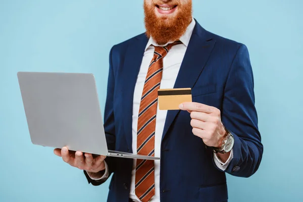 Vista recortada de sonriente hombre de negocios barbudo en traje de compras en línea con tarjeta de crédito y portátil, aislado en azul - foto de stock