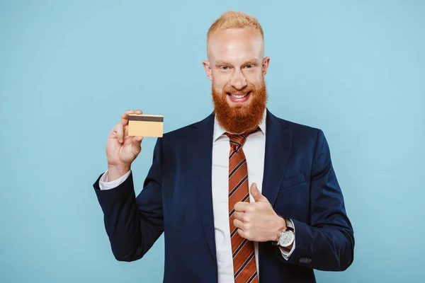 Guapo y alegre hombre de negocios sosteniendo tarjeta de crédito y mostrando el pulgar hacia arriba, aislado en azul - foto de stock