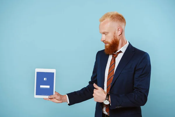 Kyiw, Ukraine - 27. August 2019: bärtiger Geschäftsmann im Anzug zeigt digitales Tablet mit Facebook-App, isoliert auf blauem Grund — Stockfoto