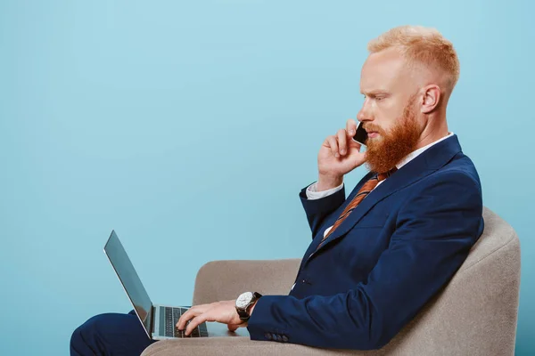 Hombre de negocios confiado hablando en el teléfono inteligente y trabajando en el ordenador portátil mientras está sentado en el sillón, aislado en azul - foto de stock