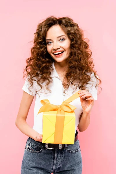 Happy curly girl holding yellow gift box, isolated on pink — Stock Photo