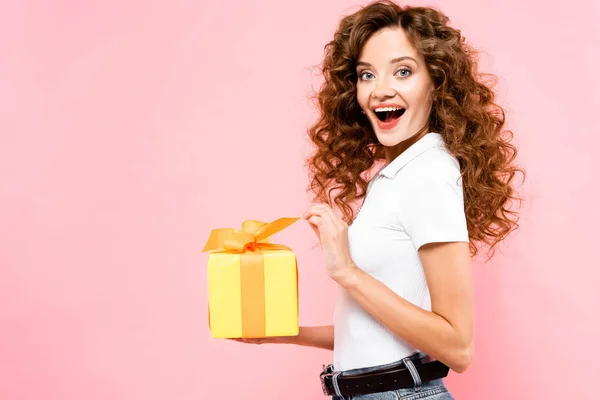 Excitada atractiva chica rizada sosteniendo caja de regalo, aislado en rosa - foto de stock