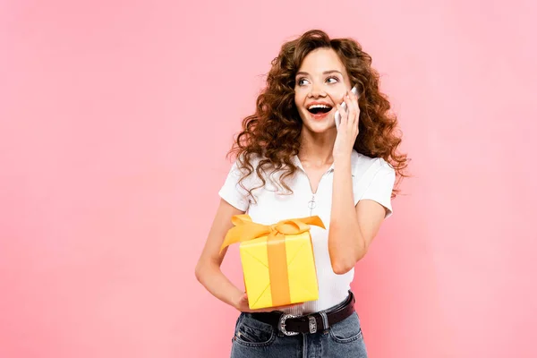 Excited curly girl holding gift box and talking on smartphone, isolated on pink — Stock Photo