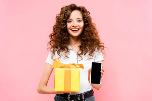 Alegre chica rizada con caja de regalo que muestra el teléfono inteligente con pantalla en blanco, aislado en rosa - foto de stock