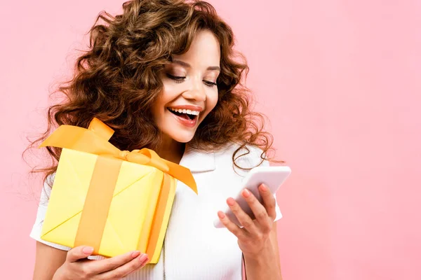 Feliz chica rizada sosteniendo caja de regalo y el uso de teléfono inteligente, aislado en rosa - foto de stock