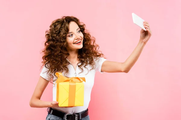 Attrayant heureux fille bouclée prendre selfie avec boîte cadeau, isolé sur rose — Photo de stock