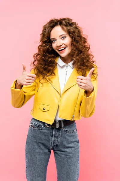 Atractiva chica rizada en chaqueta de cuero amarillo que muestra los pulgares hacia arriba, aislado en rosa - foto de stock