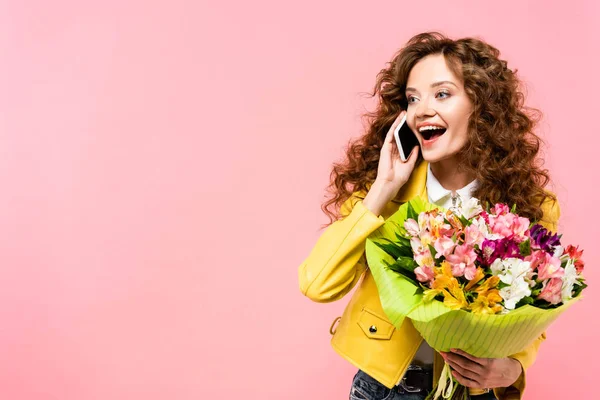 Happy curly girl with bouquet of flowers talking on smartphone, isolated on pink — Stock Photo