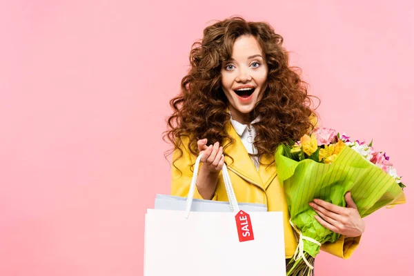 Excitada chica rizada sosteniendo flores y bolsas de compras con etiqueta de venta, aislado en rosa - foto de stock