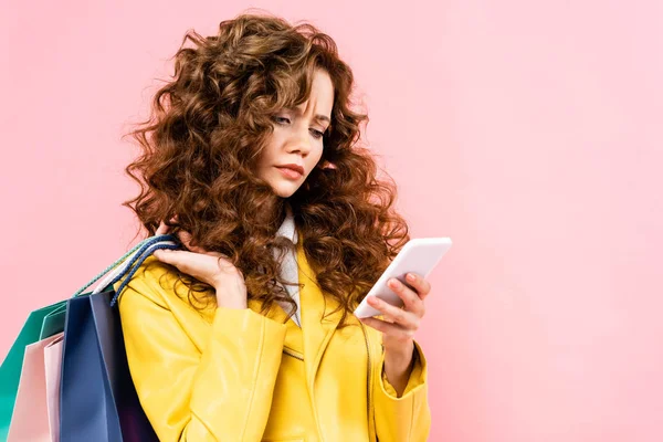 Schönes nachdenkliches Mädchen mit Smartphone und Einkaufstaschen in der Hand, isoliert auf rosa — Stockfoto