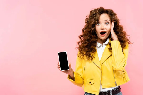 Шоковая кудрявая девушка показывает смартфон с пустым экраном, изолированный на розовый — стоковое фото