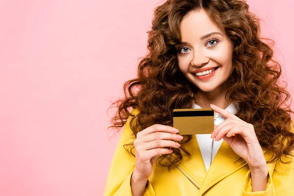 Menina encaracolado alegre atraente segurando cartão de crédito, isolado em rosa — Fotografia de Stock