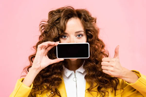 Кучерява дівчина показує великий палець вгору і смартфон з порожнім екраном, ізольована на рожевому — стокове фото