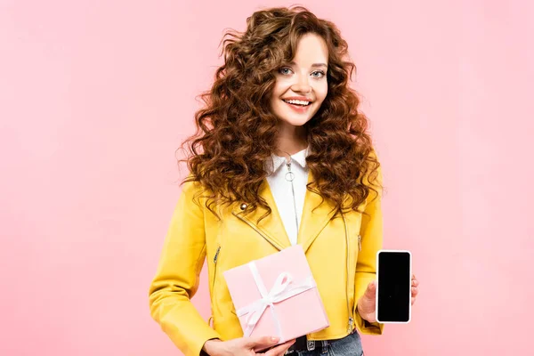 Menina encaracolado atraente mostrando presente e smartphone com tela em branco, isolado em rosa — Fotografia de Stock