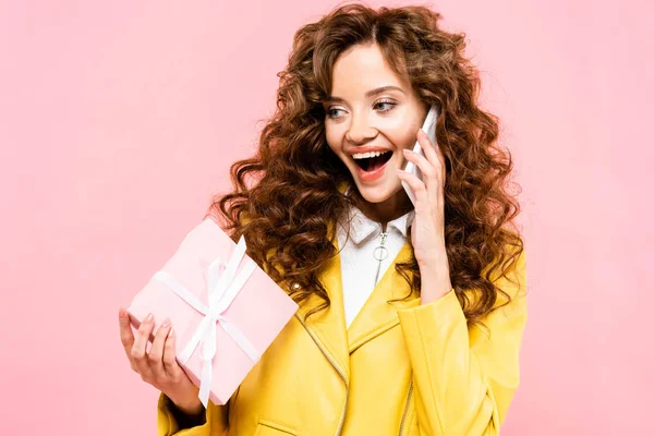 Attraente ragazza riccia parlando su smartphone e in possesso di regalo, isolato su rosa — Foto stock