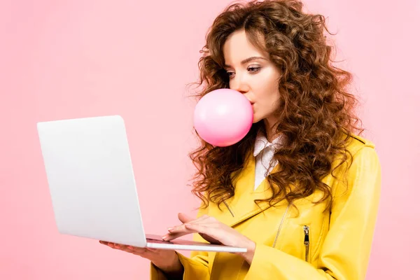 Heureuse femme bouclée avec bulle de gomme à mâcher à l'aide d'un ordinateur portable, isolé sur rose — Photo de stock