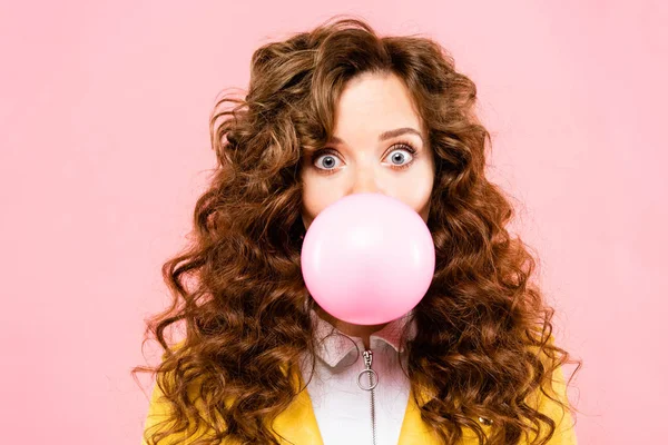 Atractiva chica rizada sorprendida con goma de mascar burbuja, aislado en rosa - foto de stock