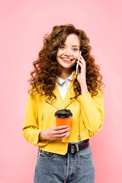 Chica alegre con taza de café desechable hablando en el teléfono inteligente, aislado en rosa - foto de stock