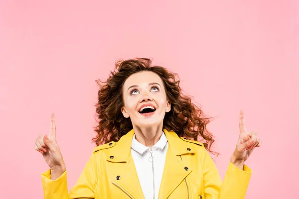 Surprised curly girl in yellow leather jacket pointing up, isolated on pink — Stock Photo