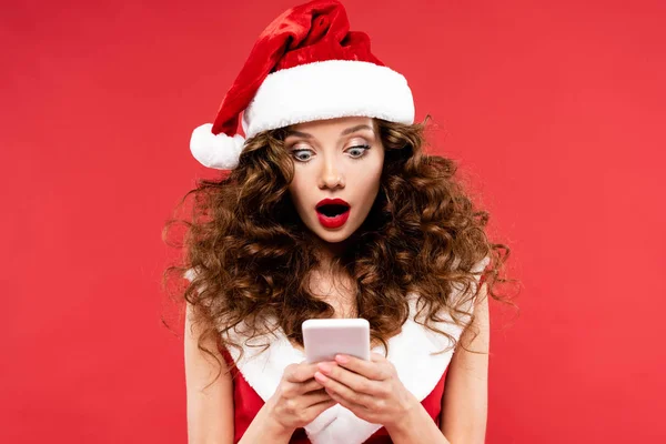 Femme choquée en costume de Père Noël à l'aide d'un smartphone, isolé sur rouge — Photo de stock