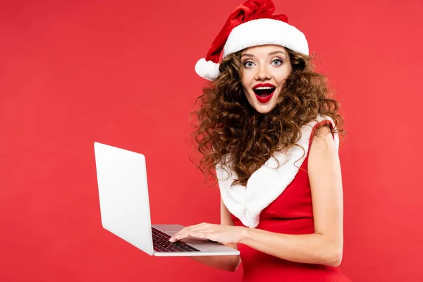 Atractiva mujer excitada en traje de santa usando el ordenador portátil, aislado en rojo - foto de stock