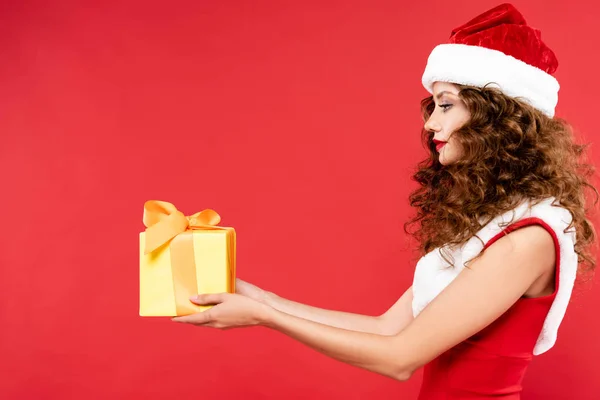 Curly girl in santa costume holding christmas gift, isolated on red — Stock Photo