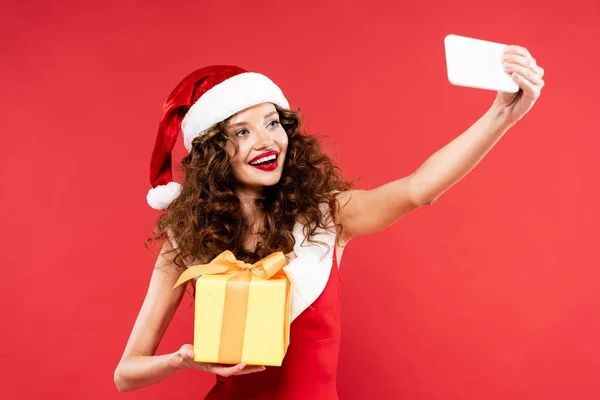 Cheerful girl in santa costume holding christmas gift while taking selfie on smartphone, isolated on red — Stock Photo