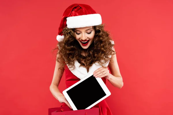 Gaie fille en costume de Père Noël tenant sac à provisions avec tablette numérique, isolé sur rouge — Photo de stock
