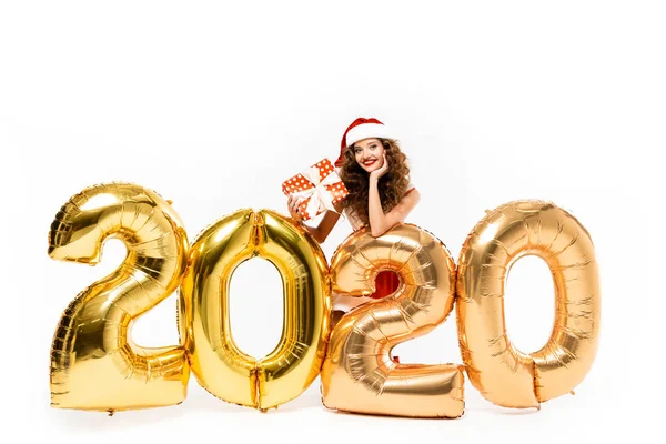 Glückliches Mädchen im Weihnachtsmannkostüm hält Geschenkbox, während sie mit goldenen Luftballons posiert 2020 Neujahr, isoliert auf weiß — Stockfoto