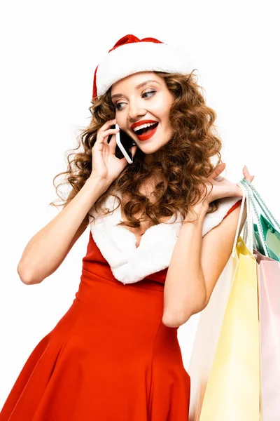 Hermosa chica rizada en traje de santa celebración de bolsas de compras y hablar en el teléfono inteligente, aislado en blanco - foto de stock