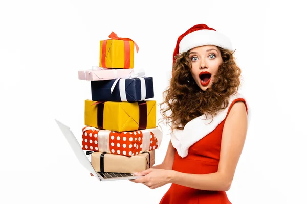 Fille choquée en costume de Père Noël tenant ordinateur portable avec pile de cadeaux, isolé sur blanc — Photo de stock