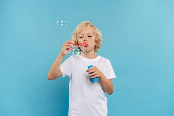 Ребенок в белой футболке, надувающий мыльные пузыри на голубом фоне — стоковое фото