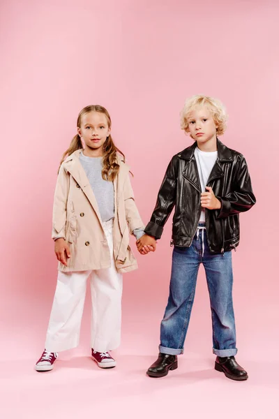 Niños con abrigo y chaqueta de cuero tomados de la mano y mirando a la cámara sobre fondo rosa - foto de stock