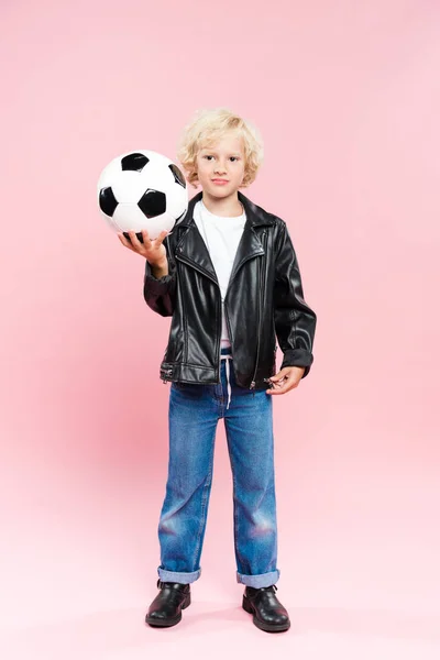 Ребенок в кожаной куртке держа футбол и глядя на камеру на розовом фоне — стоковое фото