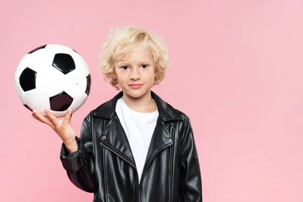 Niño en chaqueta de cuero sosteniendo el fútbol y mirando a la cámara aislada en rosa - foto de stock