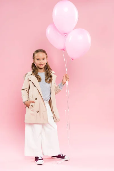 Enfant souriant en tenue d'automne avec la main dans la poche tenant des ballons sur fond rose — Photo de stock